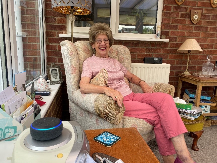 An elderly woman sitting in a decorative armchair as she smiles at an amazon Alexa.