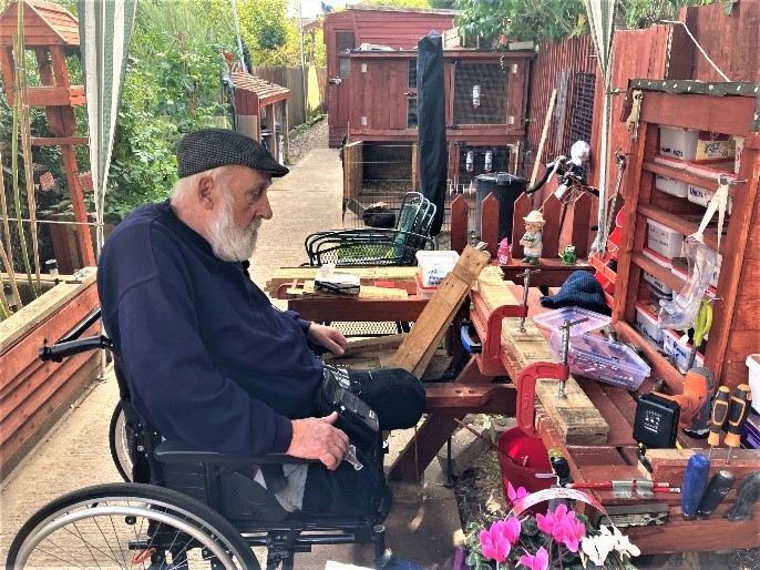 An elderly man who is using a wheelchair is focused on his worktable. Clamps are being used to hold a large piece of wood in place which he is working.