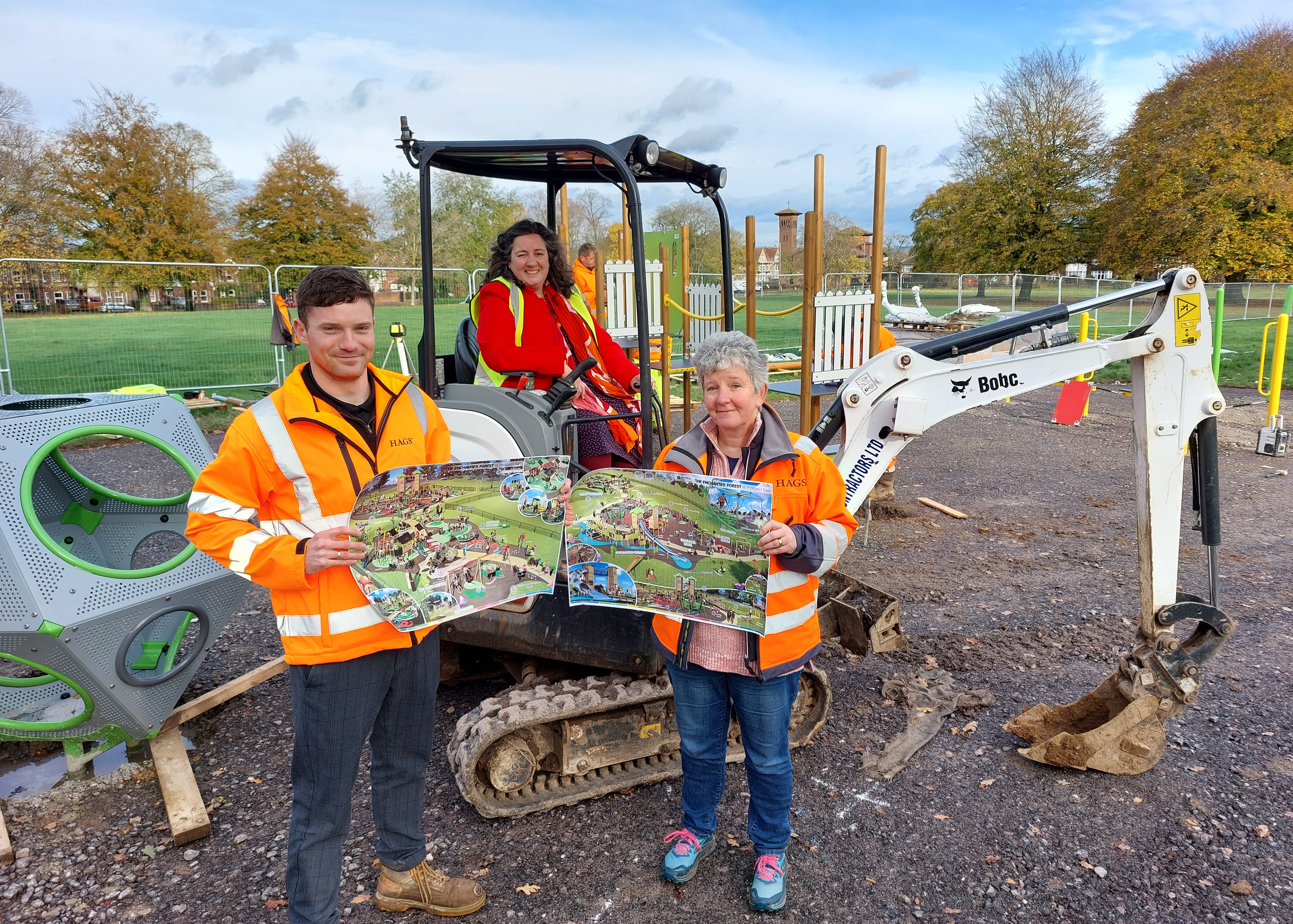 Within a playground construction workers are using equipment in the background. In the foreground two construction workers are holding up a decorative plan of what the park will look like after the construction work has finished.