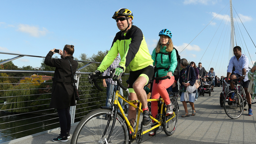 Cyclists are crossing a bridge on a tandem bike. They are looking happy as they pass pedestrians by.