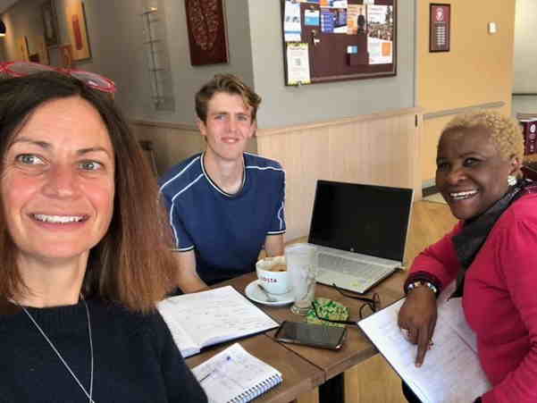 Two women and a man smiling into the camera. They are sat around a table with notebooks, a laptop and coffee.