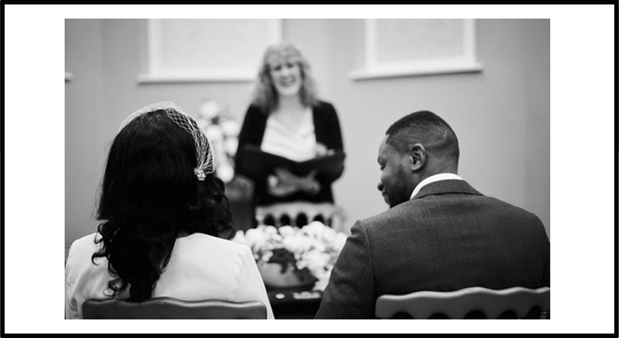 Bride and groom sat in the Ceremony Room as their ceremony is underway, with registrar smiling in the background.