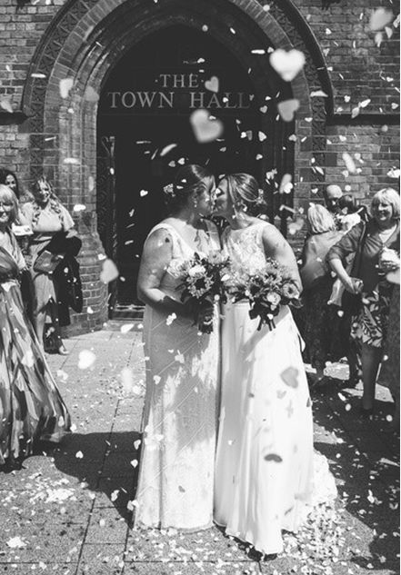 Two brides kissing outside the Town Hall, being showered with confetti.