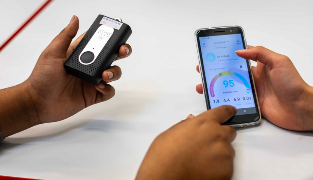 Shows three hands, the left hand of one person is holding the personal air monitor and the right hand is pointing at a phone held by another hand. The phone shows an air quality reading.