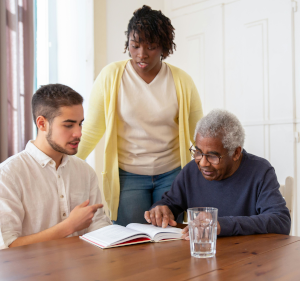 Two people sat at a table with another person stood behind, the older man (seated) is pointing at a book.