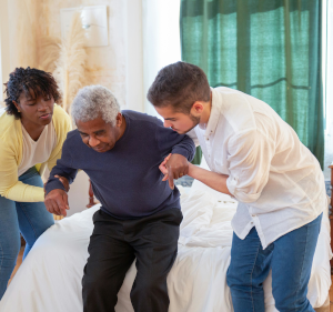 Older man being helped to stand up from a bed by a man and woman.