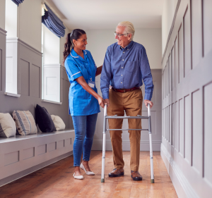 Female healthcare professional helping older man walk down a corridor using a zimmer frame.