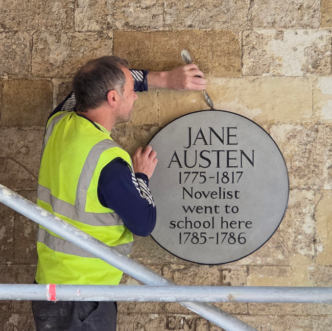 A worker in a high-visibility vest is installing a circular plaque on a stone wall. The plaque reads: 'JANE AUSTEN 1775–1817 Novelist went to school here 1785–1786.' The worker's face is blurred, and there is scaffolding in the foreground.