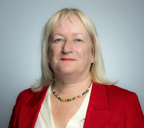 The image shows Liz Terry. It is a professional headshot of a middle-aged woman with shoulder-length blonde hair and a friendly expression. She is wearing a bright red blazer over a cream or off-white blouse, and a distinctive multi-coloured beaded necklace. The photograph appears to be taken against a neutral grey background, suggesting it's likely an official portrait or professional headshot, possibly for business or governmental purposes.