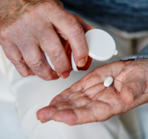 An elderly person's hands are shown. One hand is holding a white pill bottle, tilted to pour out a single white pill into the other hand. The background is blurred, focusing attention on the hands and the pill.