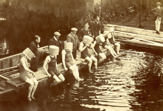 A sepia-toned photograph shows a group of me sitting on a boat in the Thames. Seven men, dressed in old-fashioned swimsuits and wearing buckets over their heads, are seated on the edge with their feet in the water. Behind them stand five men in suits are standing in the boat.