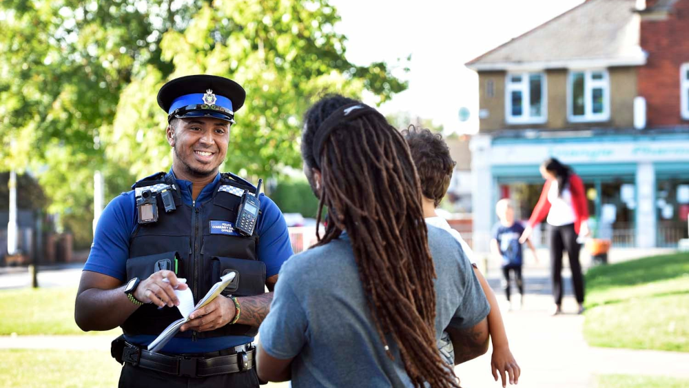 The image shows a smiling police officer in uniform having a friendly conversation with two community members on a sunny street. The officer is wearing a traditional police hat and dark vest with equipment, and appears to be engaged in positive community policing. He's speaking with a person who has long dreadlocks, with another person also present. The setting appears to be a pleasant residential or commercial area with trees, grass, and colorful shopfronts visible in the background. Other pedestrians can be seen in the distance, creating a scene of normal community life and positive police-community interaction.
