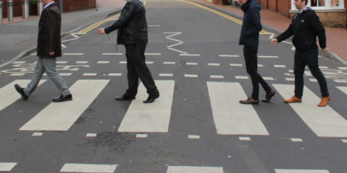  Four people in dark clothing walking across a zebra crossing (crosswalk) on a street, recreating the famous Abbey Road album cover pose by The Beatles.