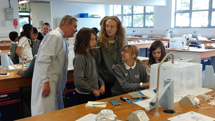 A group of secondary school students in green uniforms gather around a table in a science laboratory, examining air quality monitoring equipment with an adult instructor in a white lab coat. The students appear engaged as they learn to use the white monitoring device, with laboratory benches, large windows, and other students visible in the background of the classroom setting.