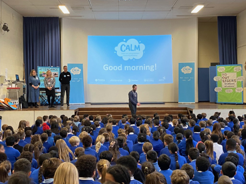 "A school assembly in a large hall with students in blue uniforms seated on the floor facing a stage. On stage, there's a large projection screen displaying 'CALM Good morning!' with the CALM logo. Several adults stand on the stage, including presenters and what appears to be school staff. Blue curtains frame the stage, and there are promotional banners visible on either side. The audience consists of dozens of primary or secondary school students.