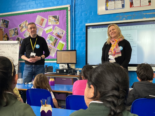 Two adults presenting to a classroom of students seated in blue chairs. A man in a dark sweater stands on the left while a blonde woman in dark clothing stands on the right, both wearing name badges or lanyards. The classroom has bright blue walls with educational displays including a purple bulletin board with photos. There's a whiteboard and desk area behind the presenters, and students can be seen from behind in the foreground listening to the presentation.