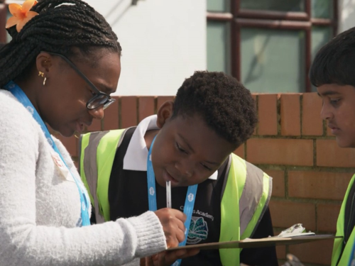 A woman with braided hair and glasses wearing a white sweater and blue lanyard leans is consulting 2 young men in a high-visibility safety vests. She is writing or working on paperwork. There is a brick wall behind them.