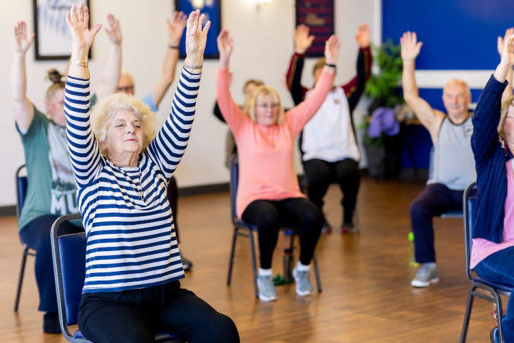 A group of older adults participate in a seated exercise class, with their arms raised high above their heads. The woman in the foreground wears a blue and white striped shirt and sits on a blue chair, while other participants in colorful clothing follow along with the movements in what appears to be a community center or gym with wooden floors.