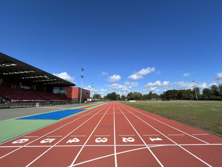 A view of the new athletics track at Palmer Park Stadium showing the red running surface with white lane markings and numbered lanes 1-8. The track has a blue section and green areas, with a covered grandstand featuring red seating on the left side. The scene is set under a bright blue sky with white clouds, surrounded by green trees and open fields.