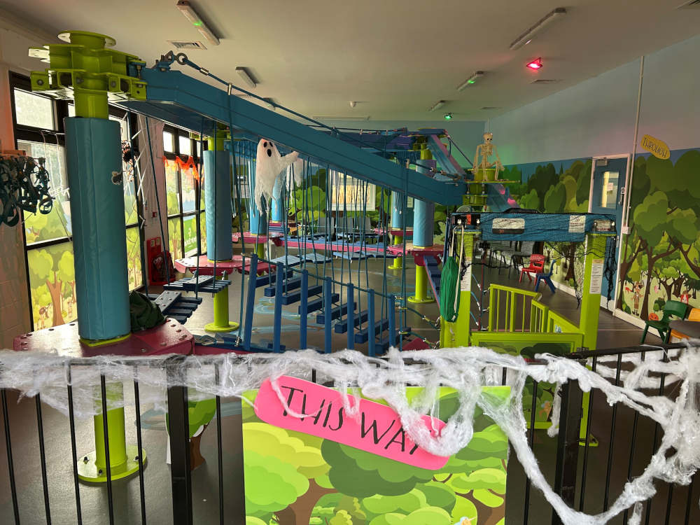 A colorful indoor adventure playground with a multi-level rope course and obstacle equipment. The space features bright lime green, teal blue, and pink accents throughout. A blue metal bridge or walkway spans across the upper level, supported by cylindrical pillars. White artificial spider webs are draped across railings in the foreground, with a pink sign reading 'IL MS WAY' (possibly 'SLIME WAY' when viewed correctly). The walls are decorated with cartoon forest murals featuring green trees and woodland scenes. Various climbing elements, suspended obstacles, and safety equipment are visible throughout the multi-story play structure.