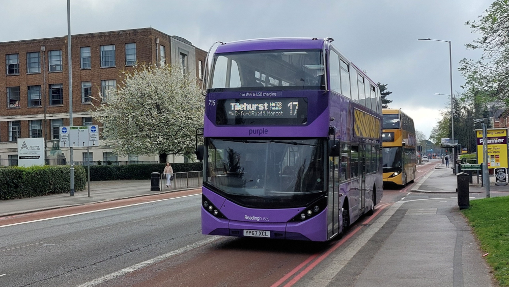 Purple double-decker Reading Buses bus number 17 to Tilehurst Water Tower traveling on an urban street. The modern bus displays 'Free WiFi & USB charging' and has license plate YP67 XCL. Behind it is a yellow double-decker bus. The scene shows a typical British street with brick buildings, trees, bus stops, and red tarmac bus lanes under an overcast sky.