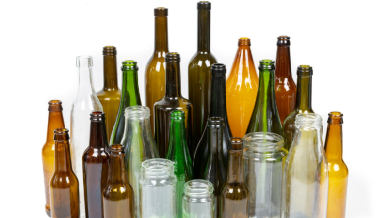 Collection of empty glass bottles and jars in various sizes, shapes and colors including clear, brown, green, and amber glass arranged against a white background. The assortment includes wine bottles, beer bottles, and glass jars of different heights, representing recyclable glass containers.