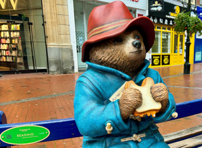 A bronze statue of Paddington Bear sitting on a blue bench in a pedestrian shopping area. The bear wears his iconic red hat and blue duffel coat, and holds a marmalade sandwich with a tag attached. A green plaque on the bench reads "READING." Behind the statue are storefronts including a bookshop, with brick paving visible on the ground.