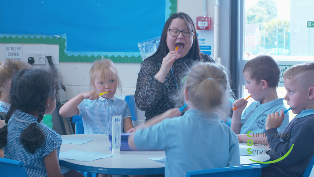 A teacher and a group of primary school children in uniform brushing their teeth together in a classroom. The children are gathered around a blue table while a female teacher with glasses demonstrates proper tooth brushing technique. The classroom has blue walls with educational displays and windows in the background. A 'Community Dental Service' logo is visible in the corner.