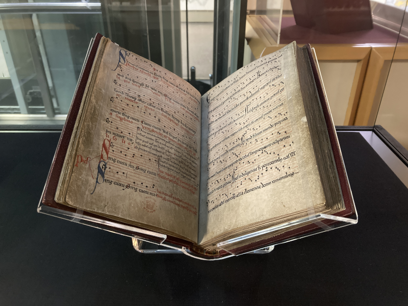 A medieval manuscript open on a display stand inside a glass museum case. The pages are aged parchment and show handwritten musical notation with lyrics in an old script. Decorative capital letters in red and blue are visible on the left-hand page. The book has a dark red cover and the pages appear worn and yellowed with age.