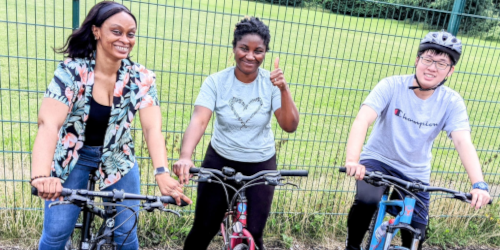 Three adults smiling and posing with bicycles outdoors near a green metal fence, taking part in a learn to ride cycling session.