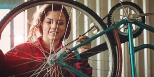 A young woman in a red jacket smiling as she works on repairing a bicycle wheel in a workshop.