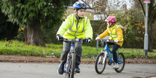 Two children wearing helmets and high-visibility jackets cycling on a road during a Bikeability cycling training session.