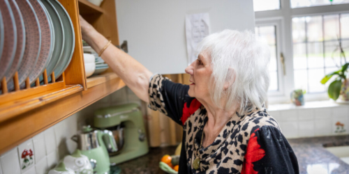 I can see an older woman with white hair reaching up to a high wooden shelf/plate rack in a kitchen to retrieve or put away plates. She's wearing a leopard print top with red accents.