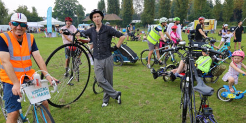 A lively outdoor cycling festival with a crowd of adults and children gathered on a grassy area with a variety of bikes, including a penny-farthing.