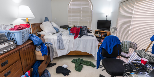 A very cluttered and untidy bedroom with clothes strewn across the bed, floor, and furniture, including a laundry basket overflowing on a dresser, a desk chair draped with clothing, and a television on a cabinet in the background.