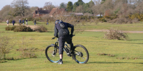 A person stands astride a mountain bike on an open grassy area, leaning forward over the handlebars. Trees, bushes and a building are visible in the background, with other people walking in the distance.