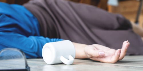 A person lying collapsed on the floor with an overturned white mug beside their outstretched hand, suggesting they have fainted or fallen, with a pair of glasses also visible on the floor nearby.