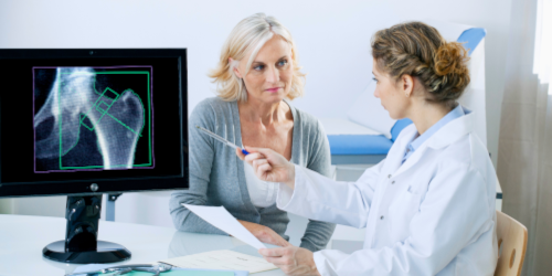 A doctor in a white coat consulting with a female patient, pointing to an X-ray image of a hip joint displayed on a monitor, in a clinical setting.