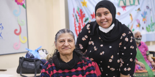 A young woman wearing a hijab smiles beside an older woman seated in front of her, with colourful artwork displayed in the background.