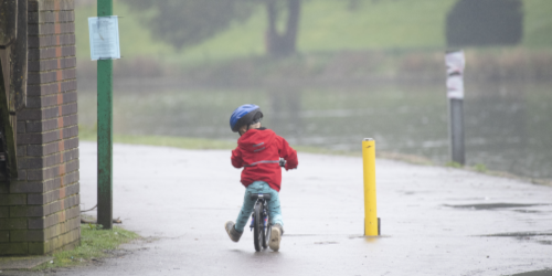 A young child wearing a blue helmet and red jacket rides a small bike along a wet path in the rain. The path runs alongside a brick building and open parkland. A yellow bollard is visible to the right.