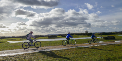 Three cyclists ride along a path through open, flat countryside under a cloudy sky. Puddles are visible on the path and surrounding grass, suggesting recent rain. Low hills can be seen in the distance.