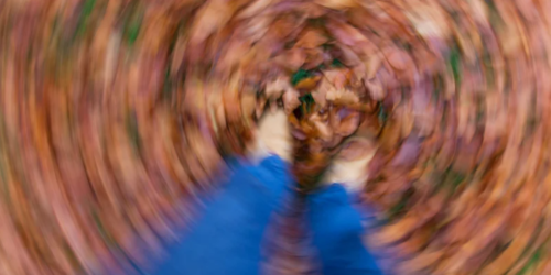 A blurred, swirling overhead shot looking down at a person's legs in blue trousers and white shoes, surrounded by autumn leaves on the ground, with a radial motion blur effect creating a dizzy, disorienting sensation.