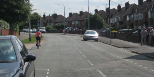 A cyclist wearing a red top rides along a residential street lined with parked cars and semi-detached houses. A silver car is pulling out from the opposite side of the road, and a pedestrian stands on the pavement to the right. The scene illustrates a typical suburban road where cyclists and drivers share the space.