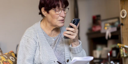 An older woman with short red hair and glasses holding a smartphone close to her face while looking at a document, seated in a home setting.