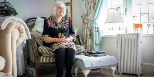 An older woman with white hair sits relaxed in a striped armchair in a cosy, eclectic living room, gazing toward a window with floral curtains.