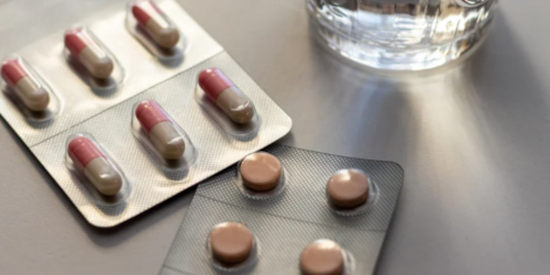 Two blister packs of medication — one containing capsules and one containing tablets — on a grey surface next to a glass of water.