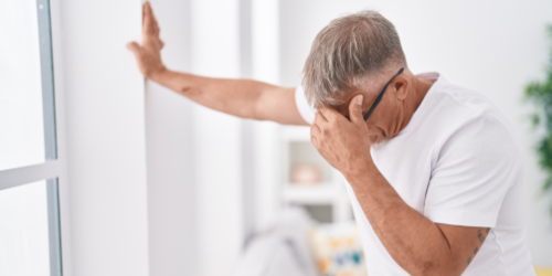 An older man leaning against a wall with one hand while holding his head, appearing dizzy or distressed.