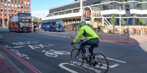 A cyclist wearing a high-visibility yellow jacket and helmet rides along an urban road. A cycle lane marking is visible on the road surface. A red double-decker bus and a coach are visible in the background, along with a multi-storey building.