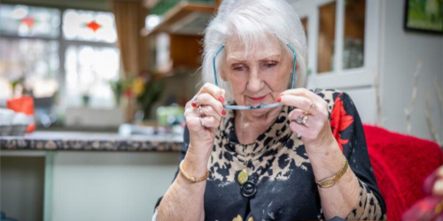 An elderly woman with white hair peering closely at her glasses while holding them up to examine them, seated indoors.