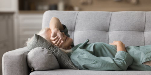 An older woman resting on a grey sofa with her arm over her face, wearing a teal top.